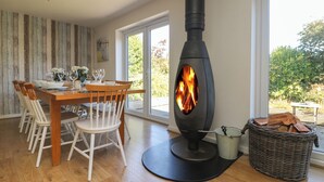 A dining area with a wooden table and chairs, a black wood stove with a fire, and a wicker basket with logs.
