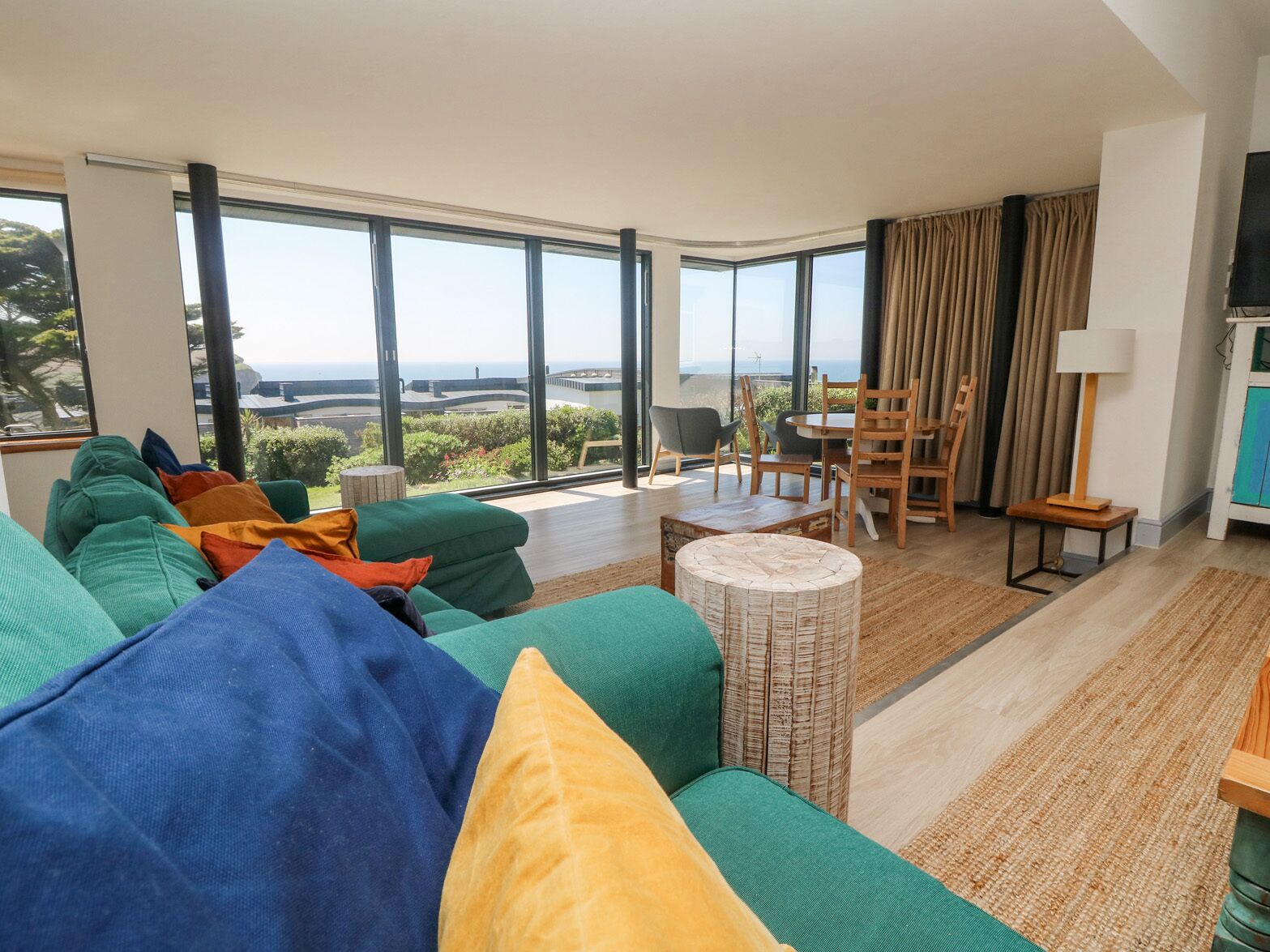 A modern living room with a large sofa, a dining table, and chairs. There is a view of the ocean through large windows.