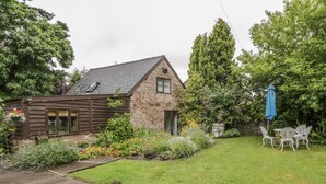 Une maison en pierre avec un toit en pente, un porche à l’avant et un petit jardin comprenant une pelouse et du mobilier de jardin.