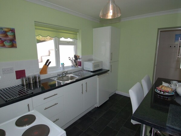 A compact kitchen with white cabinets, a black countertop, a sink, a microwave, and a dining area with a table and chairs. - Trevillas (St Austell)