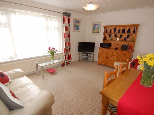 A living room with a sofa, a coffee table, a TV, a wooden cabinet with decorative items, and a window with curtains. - Trevillas (St Austell)