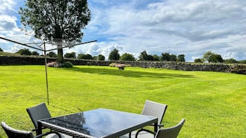 Um pátio com mesa de vidro e cadeiras de vime, rodeado por um gramado bem cuidado e paredes de pedra.