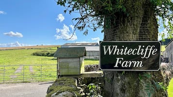 Um sinal com a inscrição 'Whitecliff Farm' preso a uma árvore, com um muro de pedra e uma estrutura de madeira ao fundo.