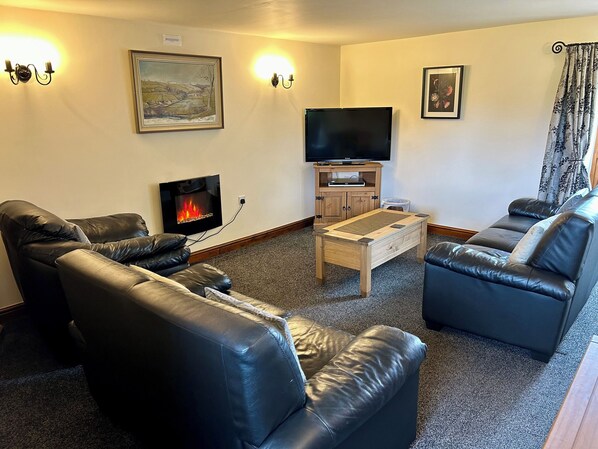 A living room with leather sofas, a flat-screen TV, a wooden cabinet, and a fireplace. - Bluebird Cottage (Buxton)