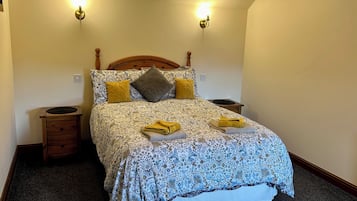 Uma cama bem arrumada com colcha estampada e almofadas, duas mesas de cabeceira e um luminário fixado na parede.