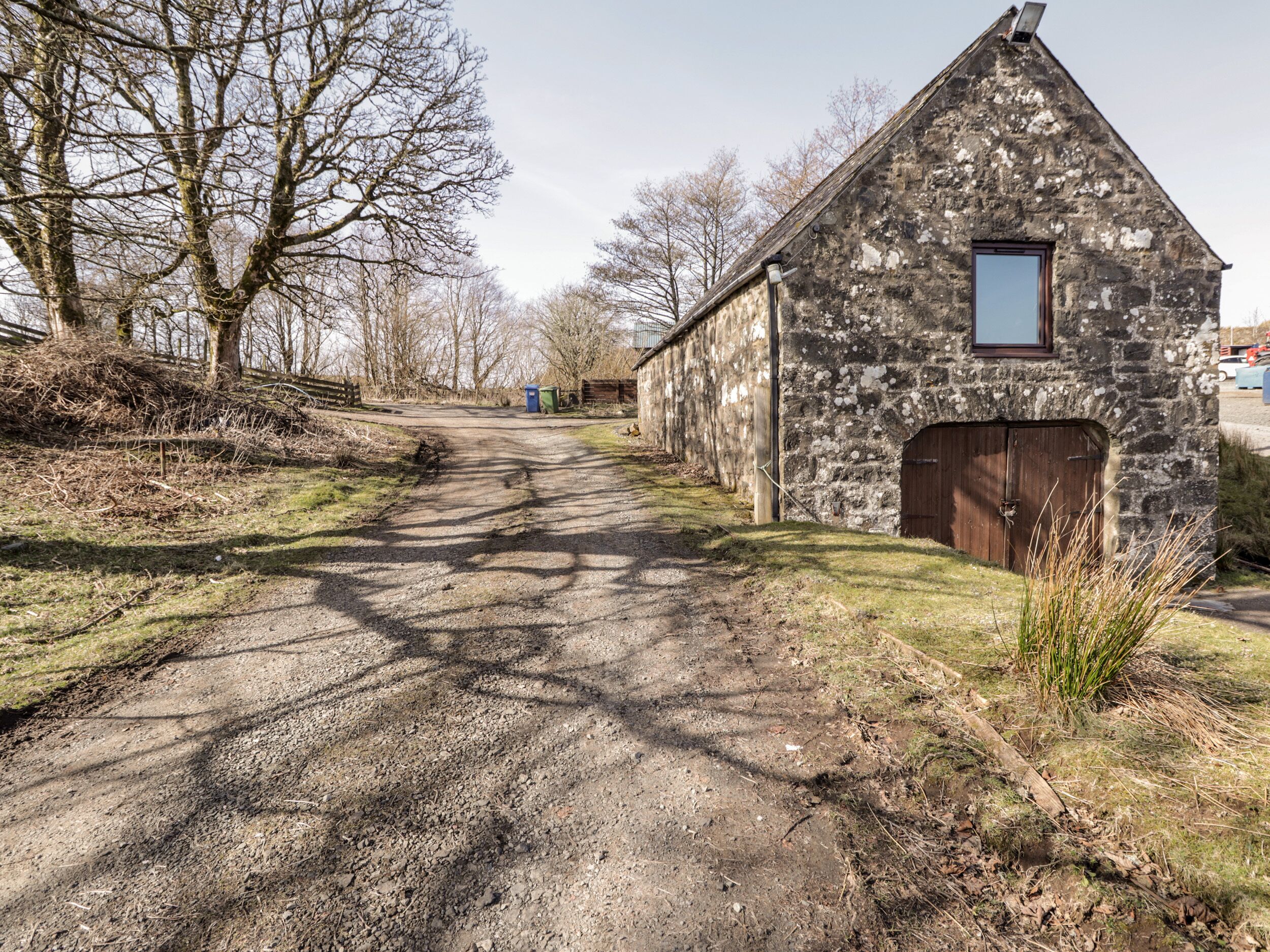 A rustic stone building with a wooden door and a small window, situated beside a gravel path with shadows cast by trees.