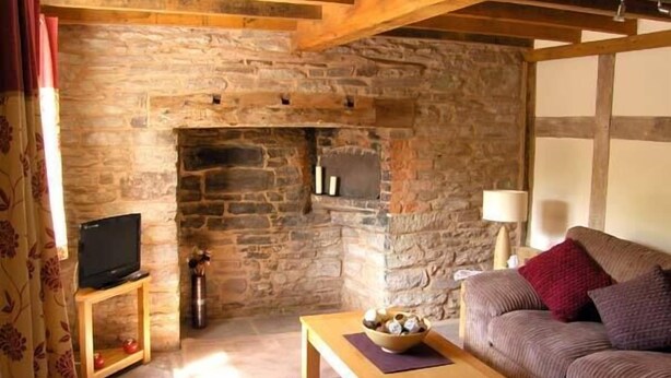 A cozy living room with a stone fireplace, a television on a wooden stand, a wooden coffee table, and a sofa with cushions. - Point Cottage (Hereford)