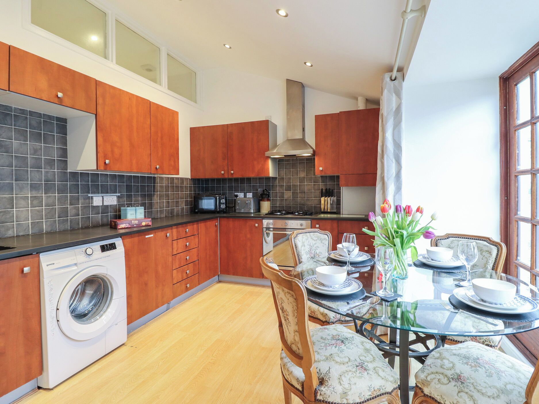 A modern kitchen with wooden cabinets, a built-in washing machine, and a dining area with a glass table and floral chairs.