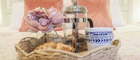 A bed with a white headboard, a pink pillow, a clear glass coffee pot, a blue and white patterned cup, a woven basket with bread, and a pink flower.