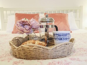 Een bed met een wit hoofdeinde, een roze kussen, een glazen koffiepot, een blauw-wit patroon beker, een rieten mand met brood, en een roze bloem.