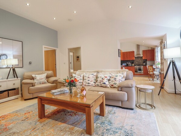 A living room with a sofa, coffee table, and a rug. There is a kitchen in the background with cabinets and a dining area.