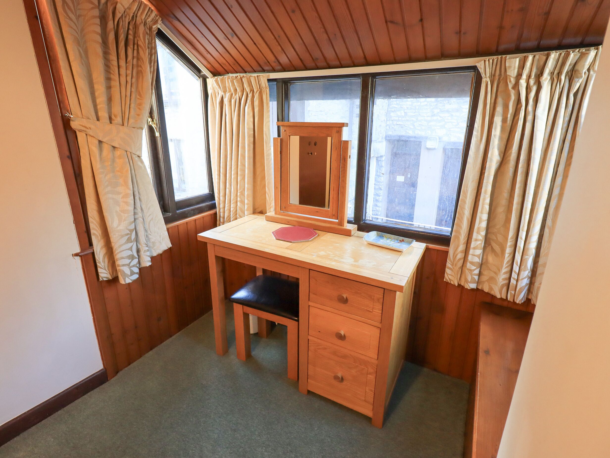 Una habitación pequeña de madera con un escritorio, una silla, un espejo y una ventana con cortinas.