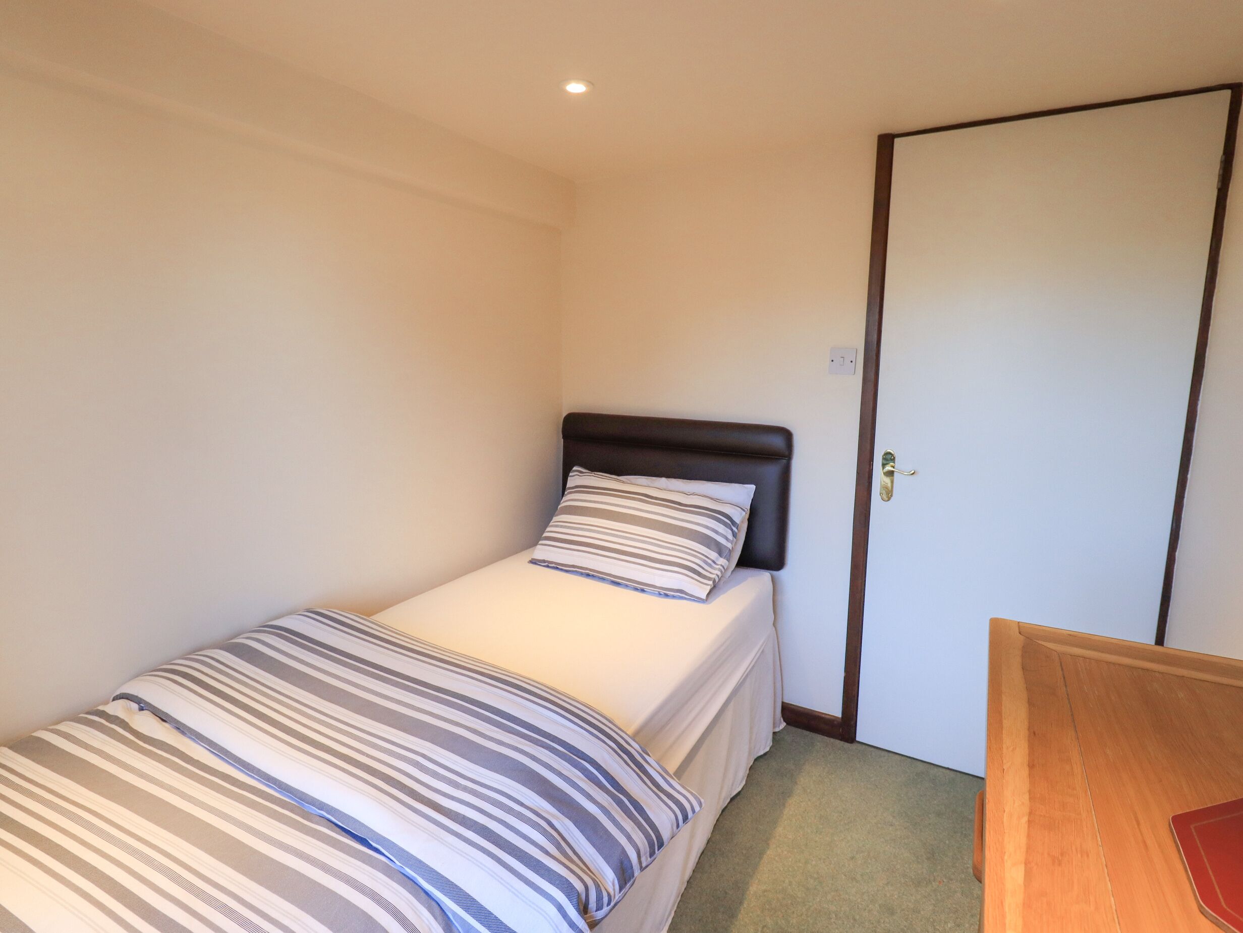 A single bed with a striped bedspread and a dark headboard in a small, well-lit room with a wooden door and a small table.
