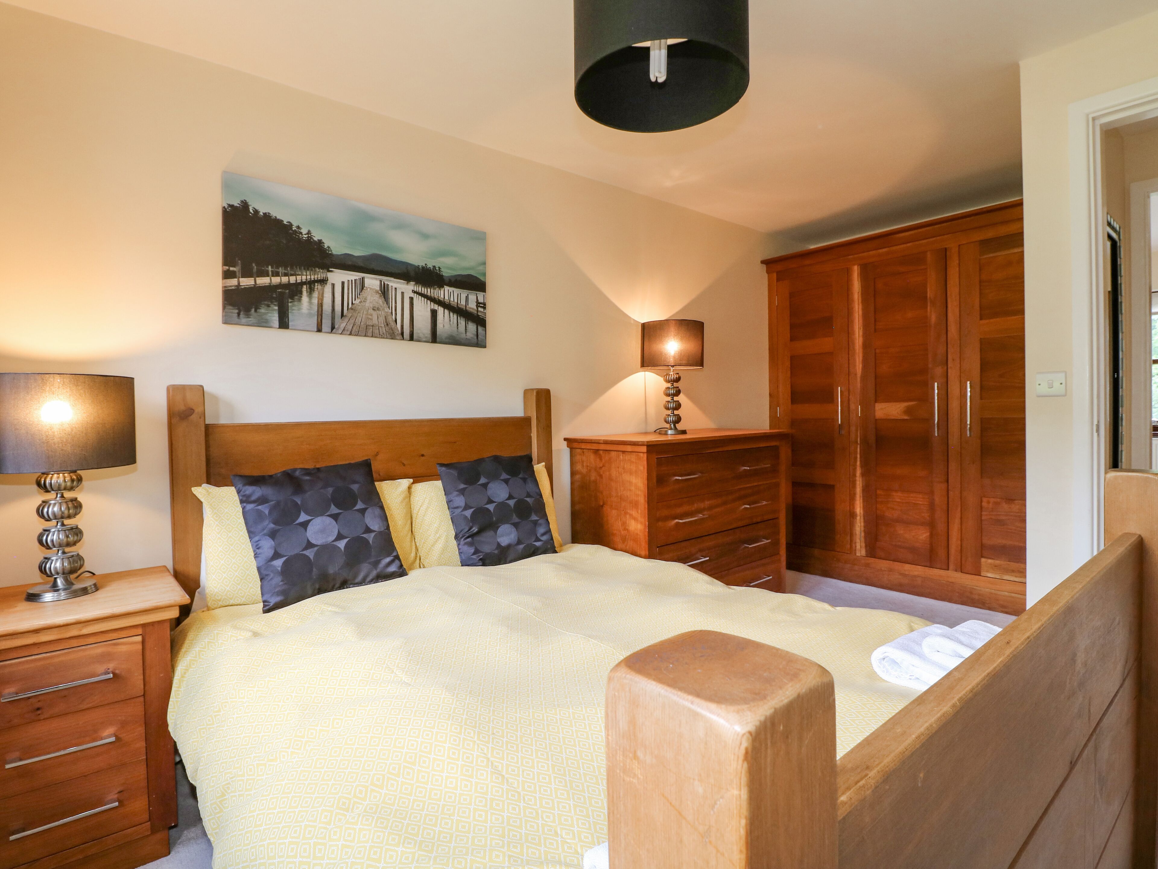 Un dormitorio con cama, cabecera de madera, mesitas de noche, lámpara, cómoda y armario.