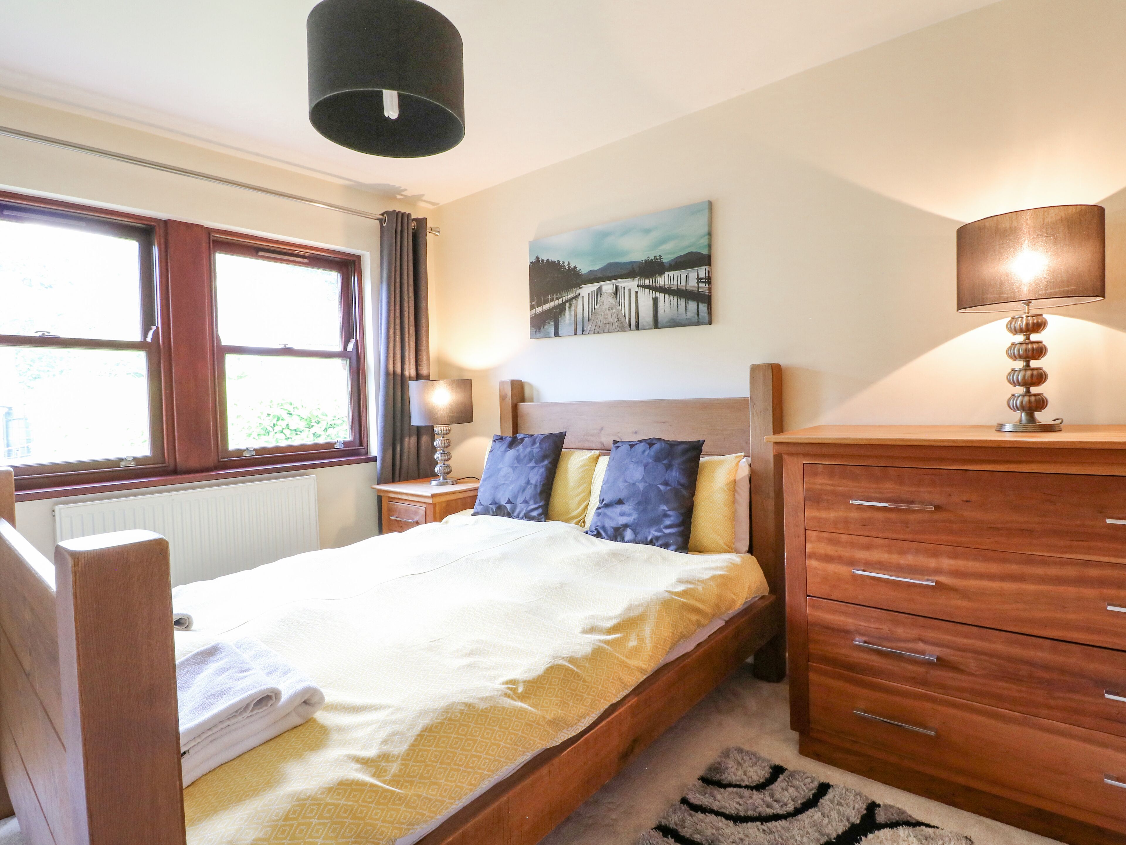 Ein Schlafzimmer mit Bett, Holz-Kopfteil, Nachttischen mit Lampen, einer Heizung, einem Fenster und einem Bild an der Wand.