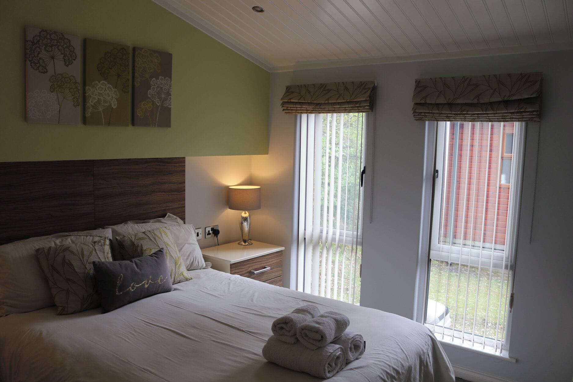 A hotel room with a bed, a bedside lamp, a window with blinds, and artwork on the wall.