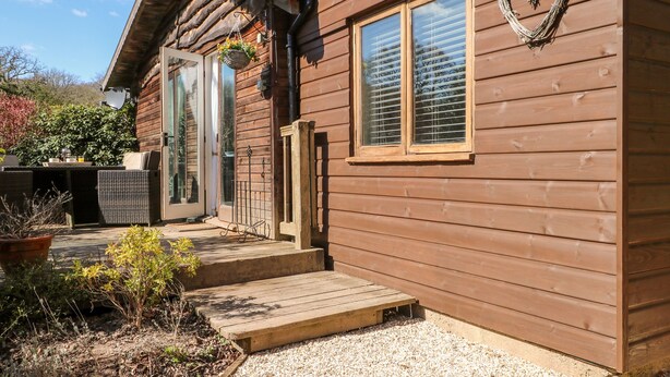 A wooden cabin with a porch, a window with blinds, and a heart-shaped decoration on the wall. - Woodmancote Lodge (Haslemere)