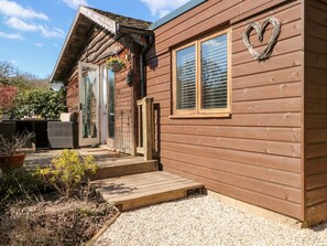 A wooden cabin with a porch, a window with blinds, and a heart-shaped decoration on the wall. - Woodmancote Lodge (Haslemere)