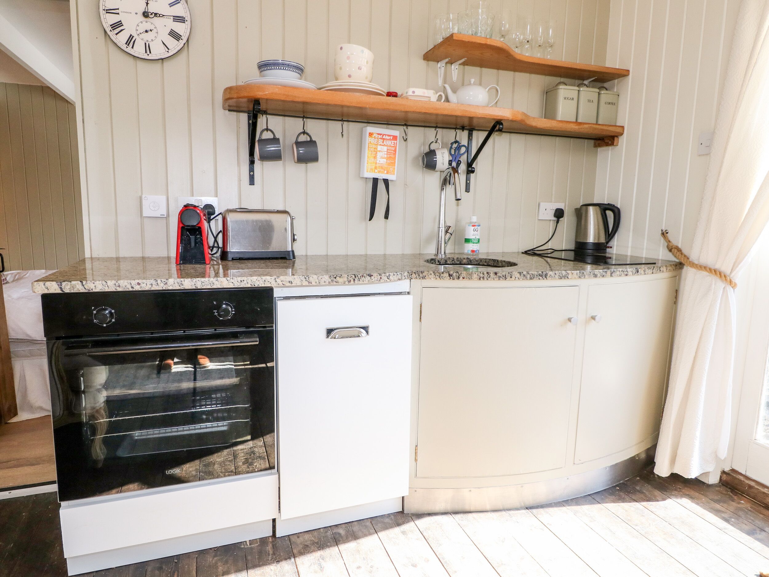 A compact kitchen with a built-in oven, a toaster, and a sink. There are shelves above the counter holding dishes and a kettle.