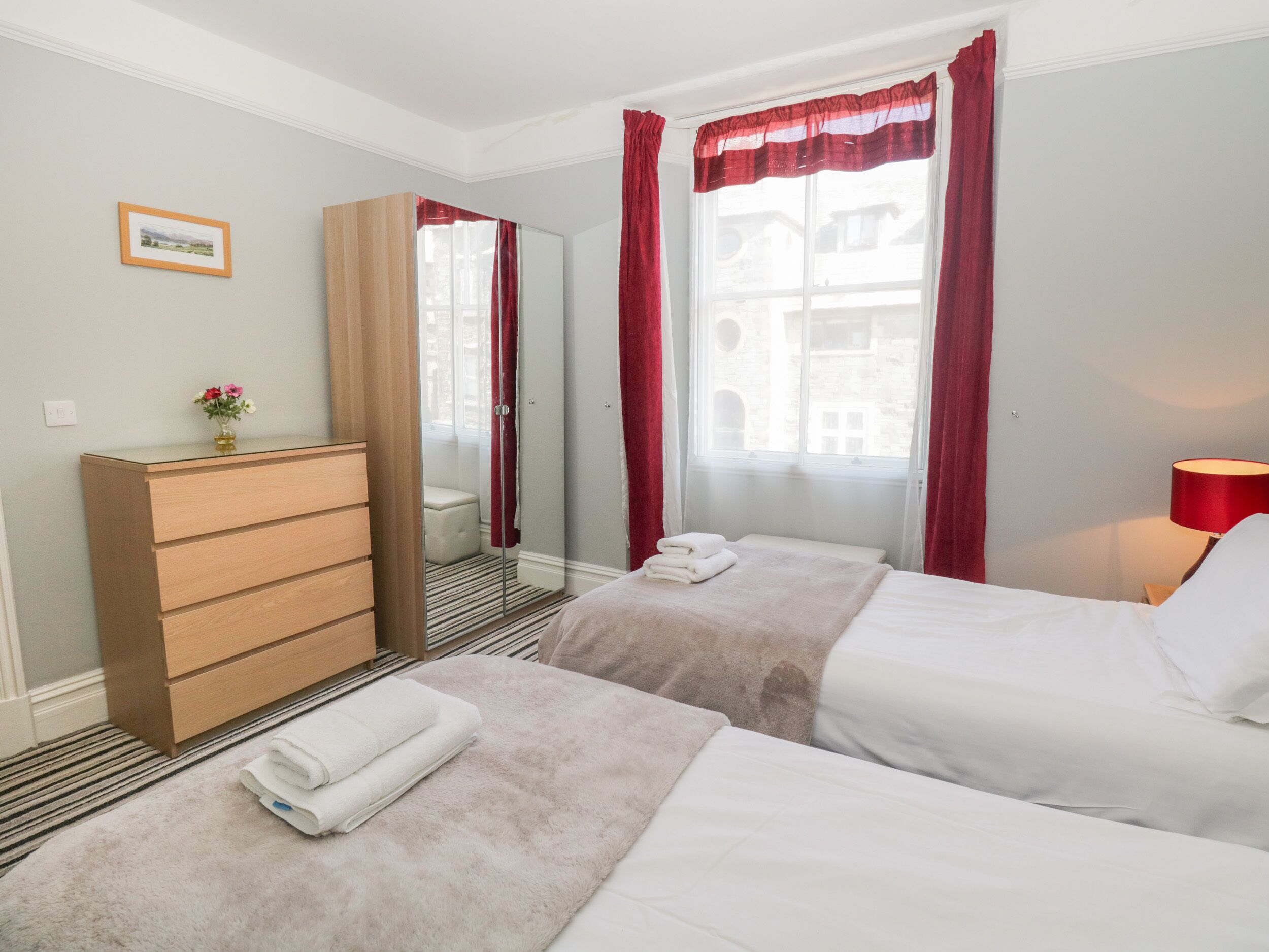 Ein Hotelzimmer mit zwei Betten, einem Holzschrank, einem Spiegel und einem Fenster mit roten Vorhängen.