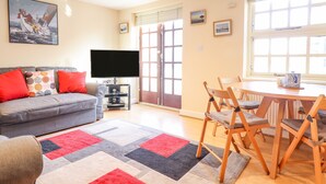 A living room with a grey sofa, red cushions, a flat-screen TV, a coffee table, and a dining area with wooden chairs. - Taliesin (Porthmadog)
