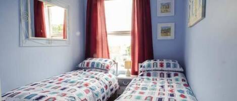 A small, well-lit bedroom with two single beds, a window with red curtains, and a mirror on the wall.