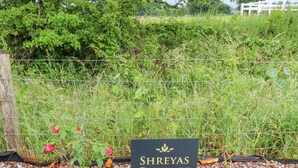 A sign for Shreyas Cottage, surrounded by a gravel path and a wire fence with greenery in the background. - Sunnyside Shreyas (Blackburn)