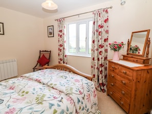 A bedroom with a bed, a wooden dresser, a chair, a window with floral curtains, and a radiator.