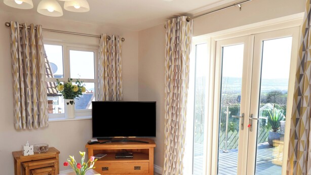 A living room with a TV on a wooden stand, a small table with a vase, and a sliding glass door leading to a balcony. - Heather Croft (Whitby)