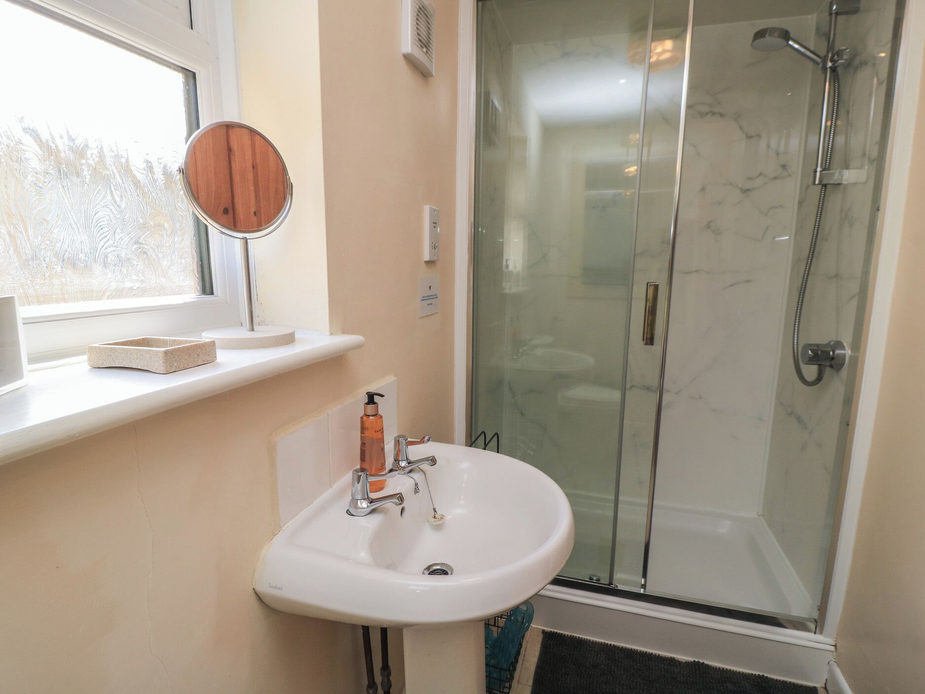 A bathroom with a white sink, a mirror, a glass shower enclosure, and a window with frosted glass.