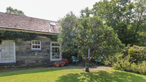 Une maison en pierre avec un toit en tuiles, un petit jardin avec une pelouse et deux chaises bleues.