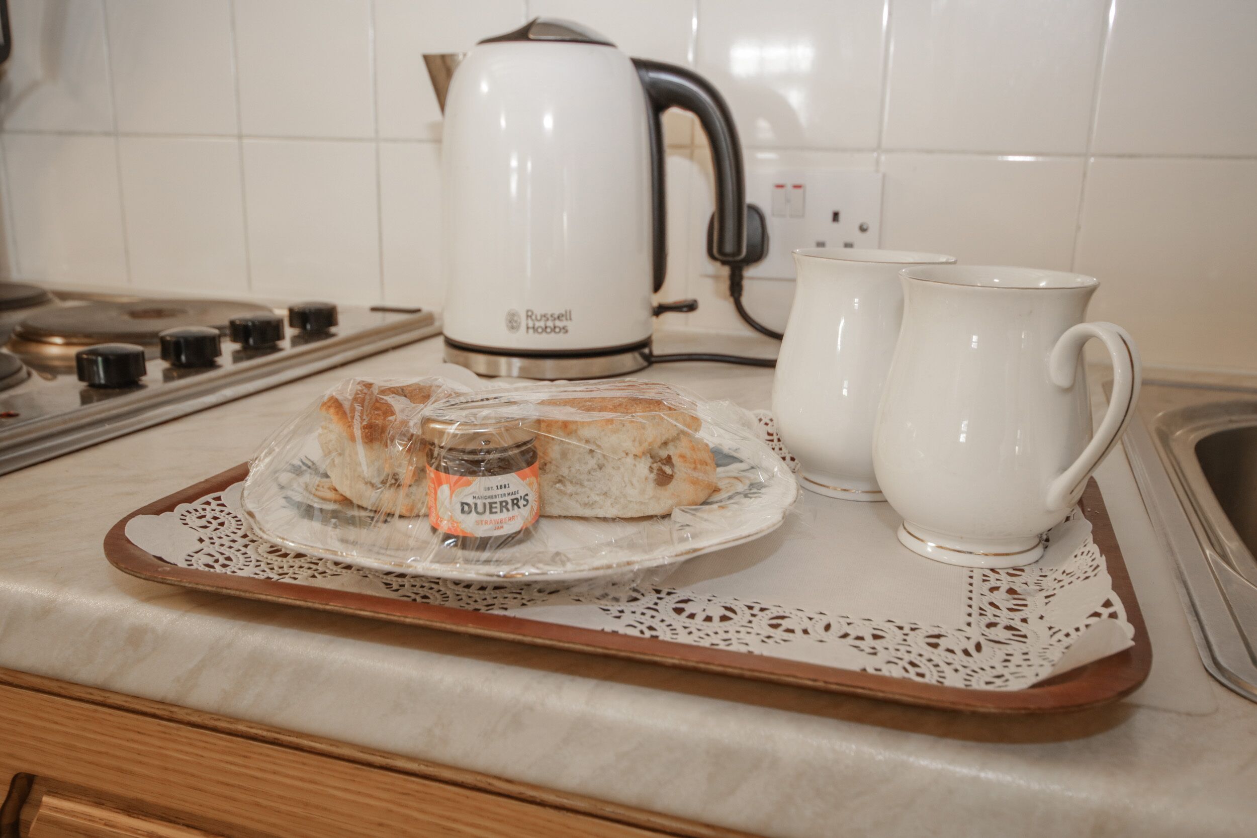 Una encimera de cocina con una bandeja que contiene un sándwich, un frasco de mermelada y dos tazas blancas.