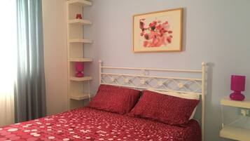 Quarto com cama, cabeceira branca, colcha vermelha com bolinhas, travesseiro rosa, prateleira fixada na parede com uma planta e uma moldura com imagem.