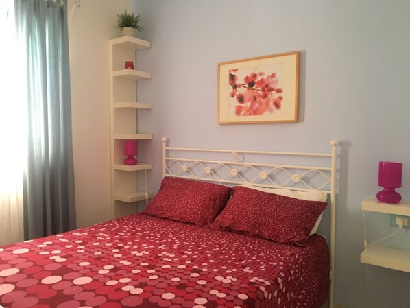 A bedroom with a bed, a white headboard, a red polka dot bedspread, a pink pillow, a wall-mounted shelf with a plant, and a framed picture. - B&B Masseria Liberatore (Mattinata)