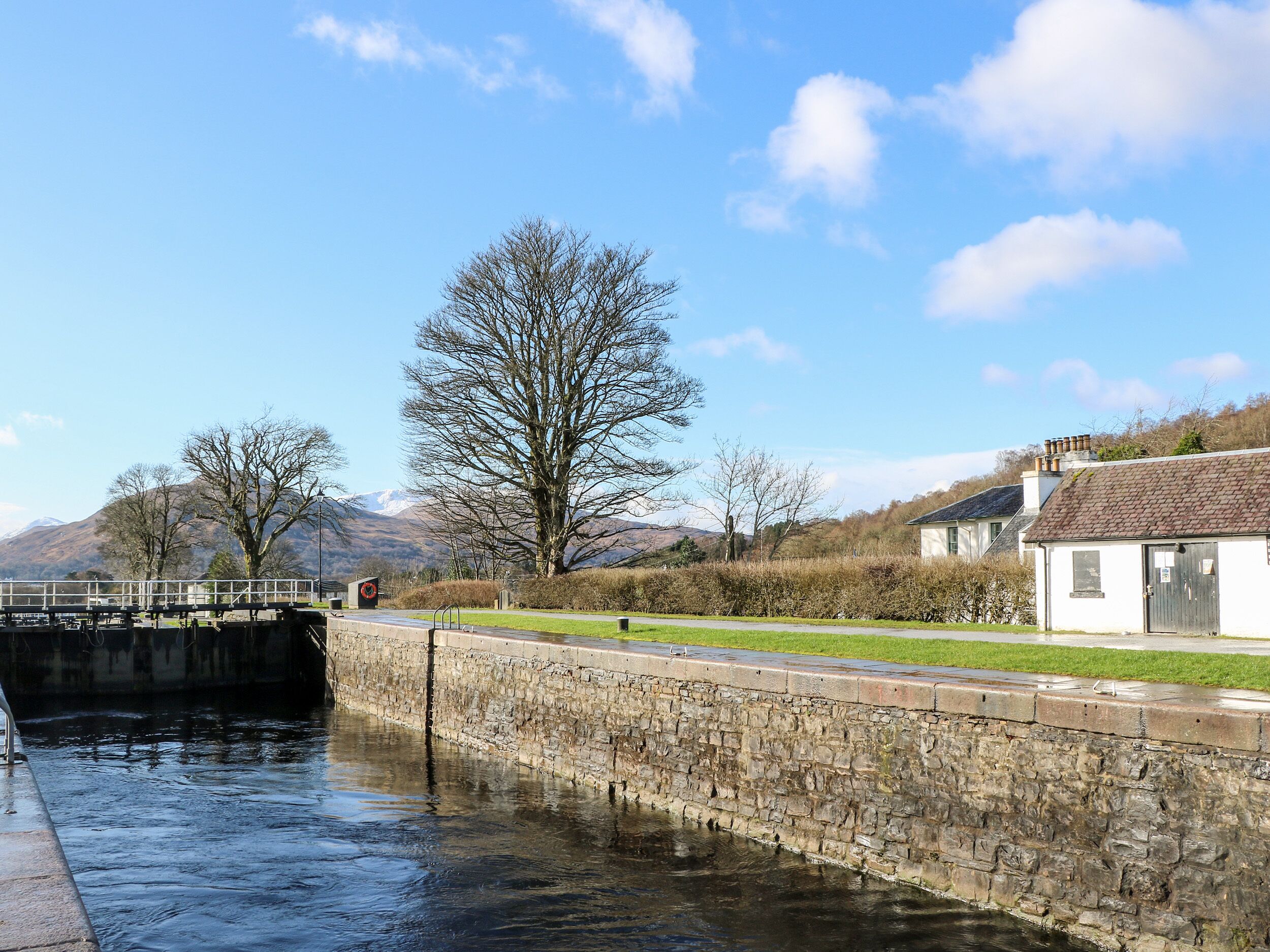 A stone lock gate by a calm river, a white building with a chimney, and a clear blue sky.