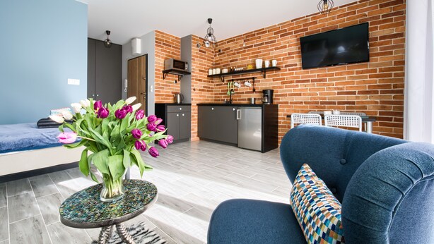 A modern living room with a blue sofa, a small round table with a vase of flowers, and a kitchen area with a brick wall and a flat-screen TV. - Roztocka Loft Apartment (Warsaw)
