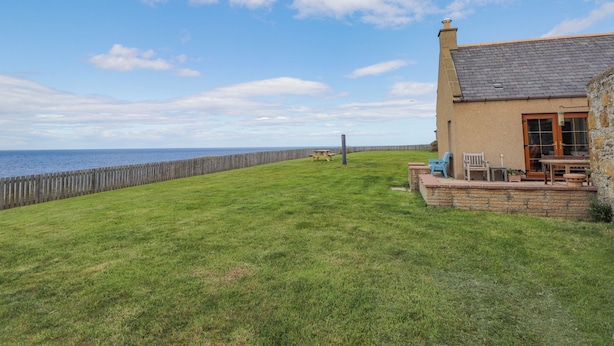 A house with a stone wall, a grassy yard, and a view of the sea. - Braehead Croft (Buckie)