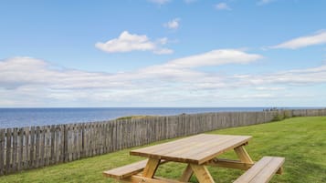 A wooden picnic table with benches in a grassy area with a wooden fence and a view of the sea.