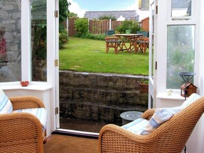 A room with wicker furniture, a stone wall, and a view of a garden with a table and chairs. - Pen y Parc (Rhyl)