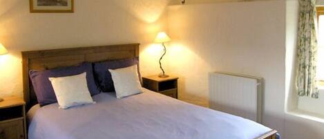 A bedroom with a wooden bed frame, white bedding, a nightstand with a lamp, a radiator, and a framed picture on the wall.