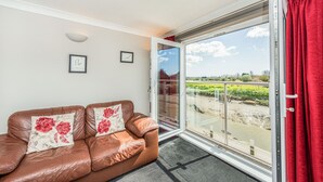 A brown leather sofa with floral cushions, a glass door leading to a balcony with a view of a river and greenery. - 16 The Boathouse (Rye)