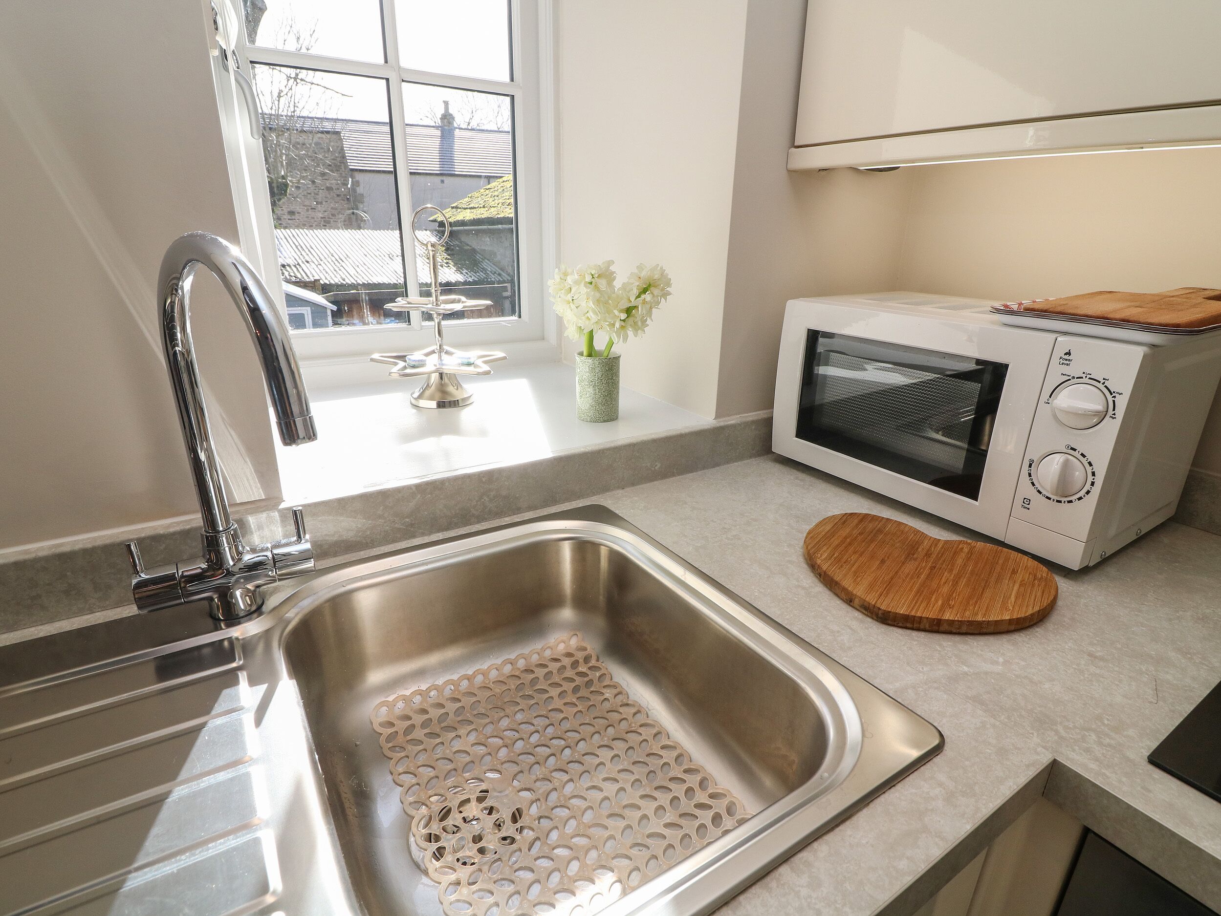 A kitchen with a stainless steel sink, a white microwave, and a window with a view of a house.