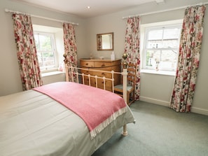 A bedroom with a bed, dresser, chair, and windows with floral curtains. - Bill's Place (Leyburn)
