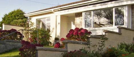 Ein zweistöckiges Haus mit Veranda, ein kleiner Garten mit verschiedenen Pflanzen und Blumen, und ein klarer Himmel.