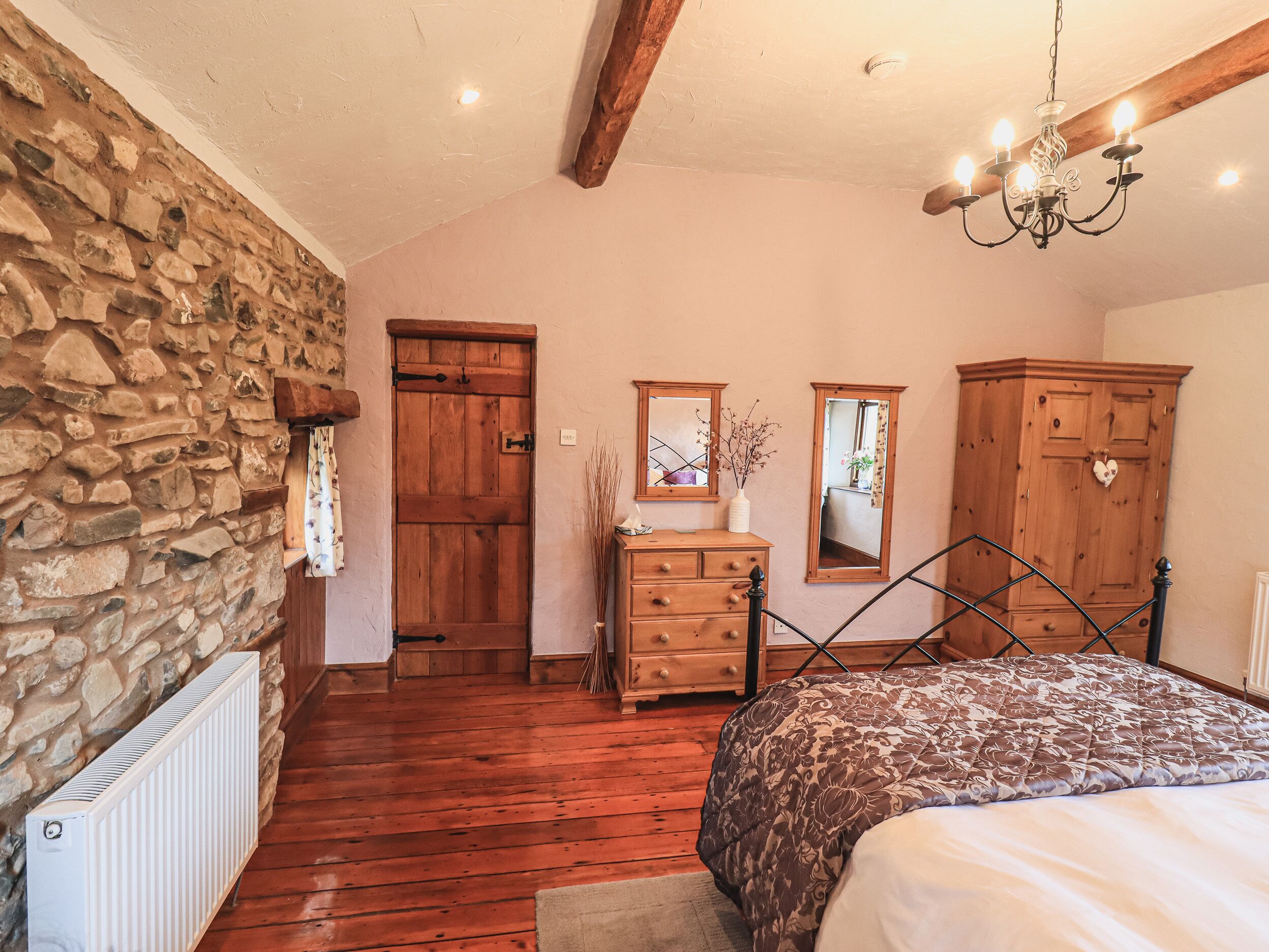 A bedroom with a stone wall, wooden door, wooden dresser, two mirrors, a chandelier, and a bed with a patterned bedspread.