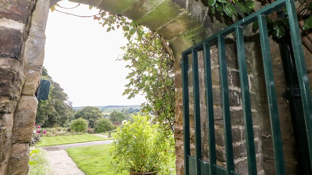Un arc en pierre mène à un jardin où se trouve un sentier, un pot de fleurs et une porte en métal.