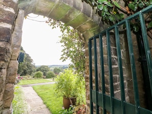 Um arco de pedra que dá acesso a um jardim com um caminho, um vaso de planta e um portão de metal.
