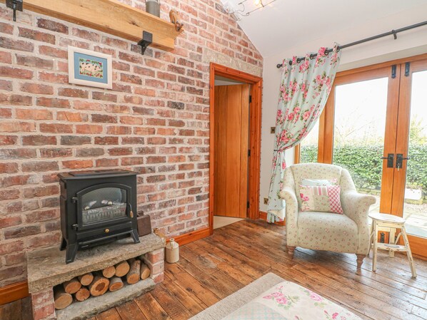 A cozy room with a brick fireplace, a floral armchair, and a wooden floor. - Maltkiln Cottage At Crook Hall Farm (Ormskirk)