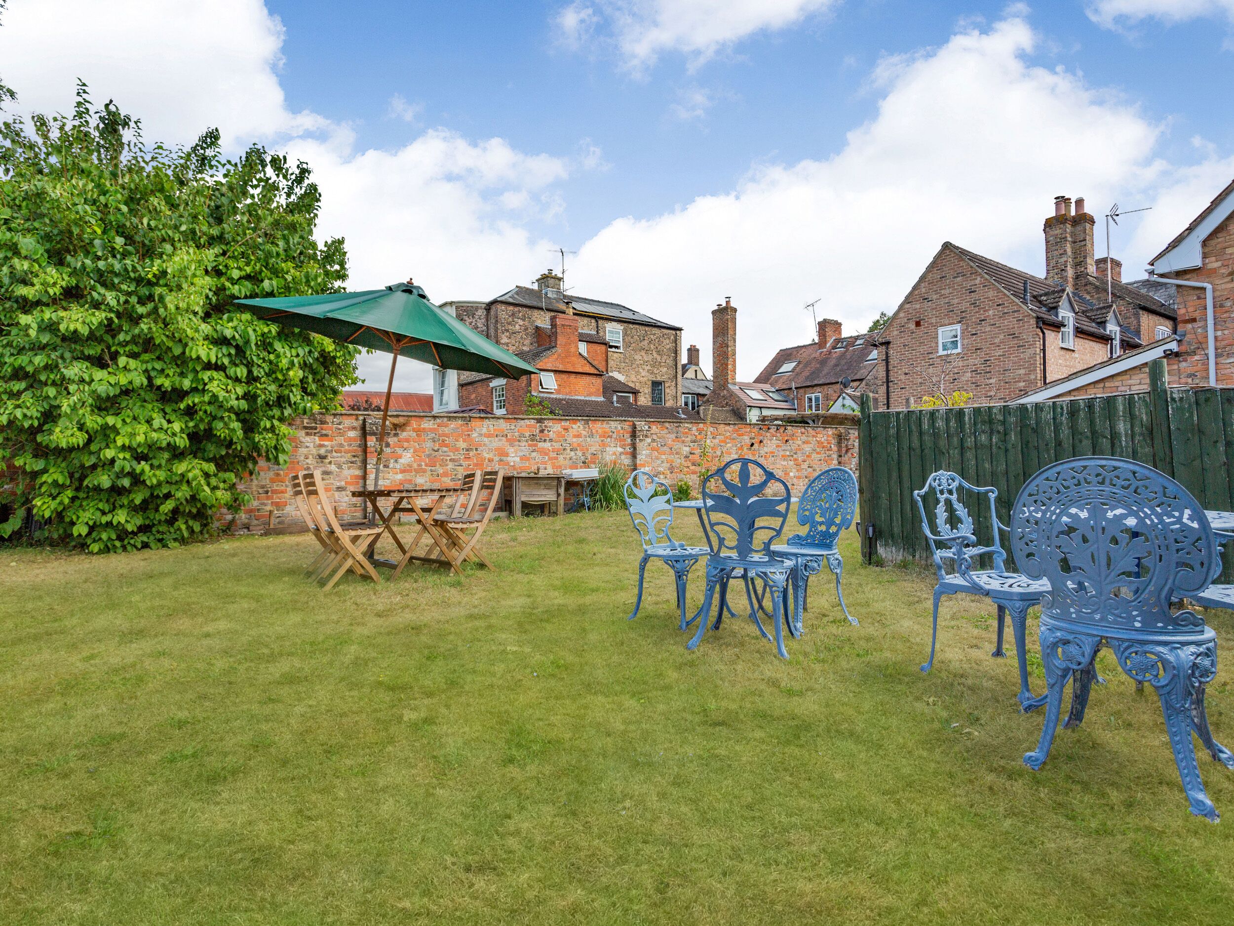 A backyard with a green umbrella, a wooden table, and blue chairs.