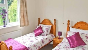 A bedroom with two wooden beds, each with a floral bedspread and purple pillows. There is a window with a view of greenery outside. - Matlock Green (Matlock)